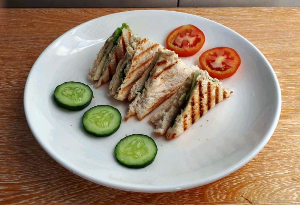 A Filipino style chicken sandwich spread  that is sliced into 4 pieces served with slices of cucumber and tomatoes