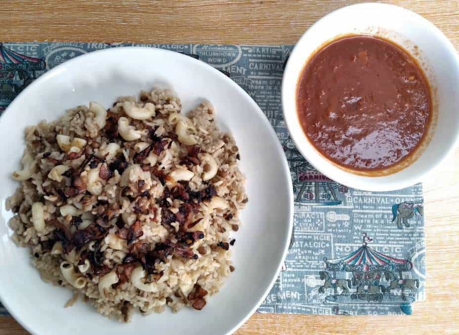 Egyptian Koshari Rice mixed with brown lentils, macaroni pasta and garnished with fried brown onion in a plate and served with tomato sauce