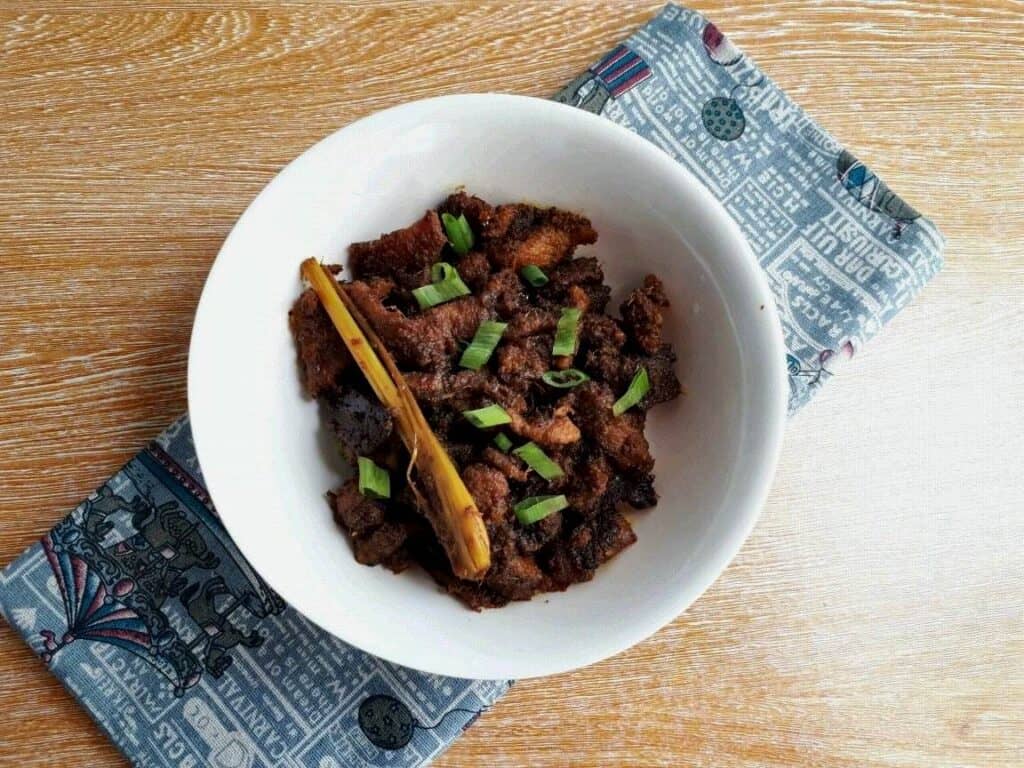 Beef Rendang recipe mixed with chopped boneless beef steak, spices & herbs, garnished with spring onion with lemongrass stalk in a plate bowl.