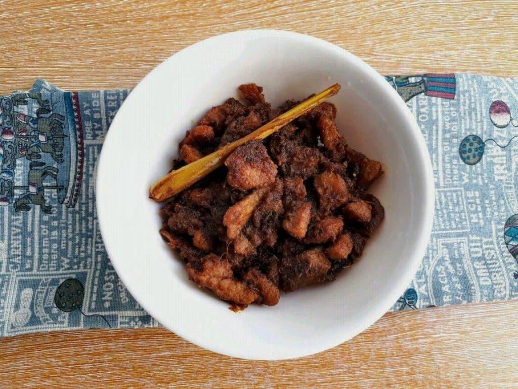 Beef Rendang recipe mixed with chopped boneless beef steak, spices & herbs, with lemongrass stalk in a plate bowl.