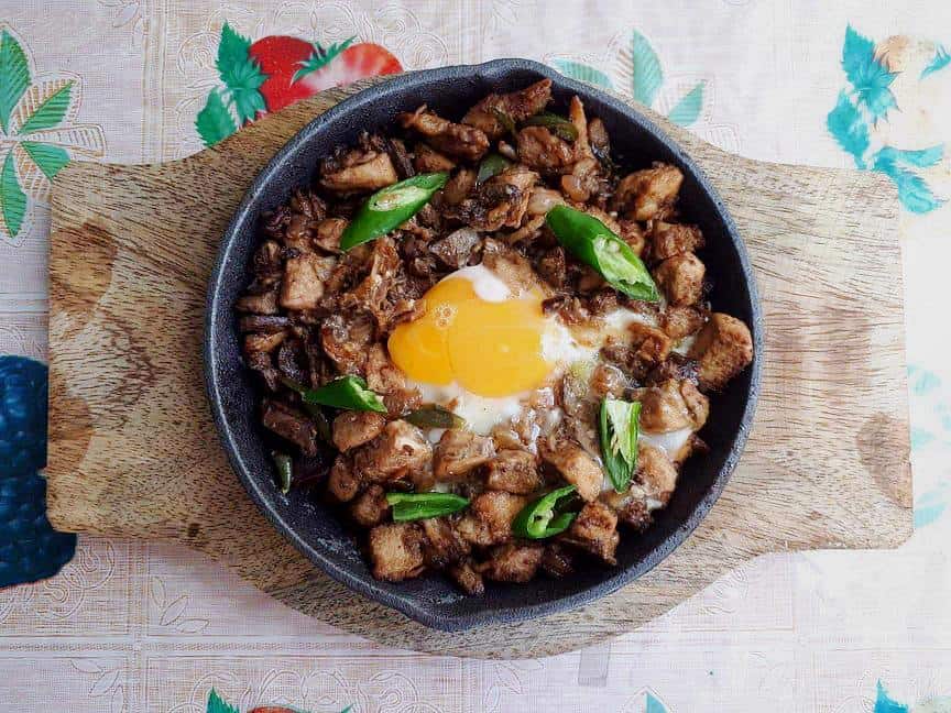 A filipino chicken sisig that is mixed with chicken liver & breast and green chilies, and topped with egg serve in a caster iron plate.