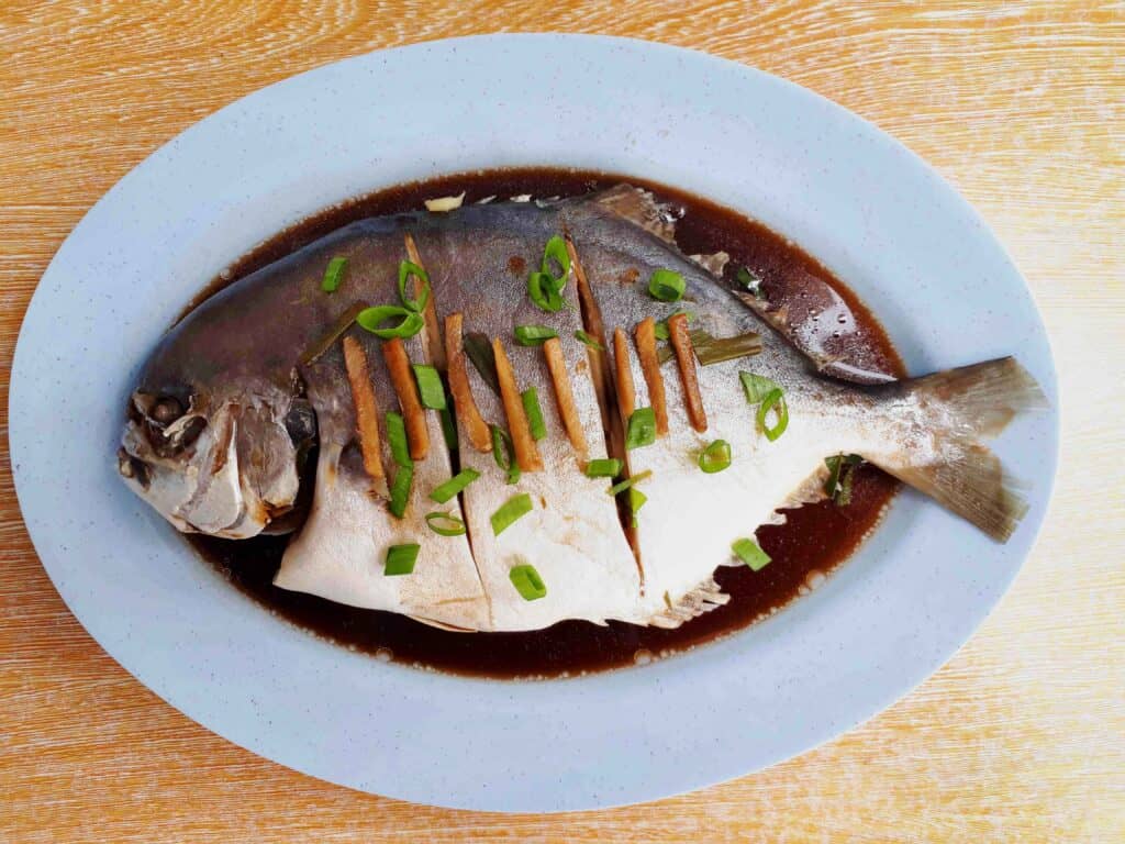 Steamed Pompano Recipe that is poured in seasoned soy sauce and garnished with chopped spring onion leaves and ginger.