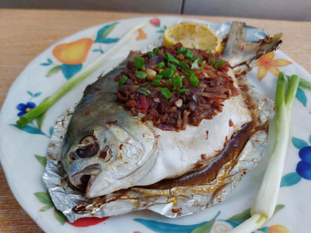 Whole baked pompano fish topped with onion-tomato soy sauce mixture with 2 onion leaves and 1 slice of lemon served on a plate