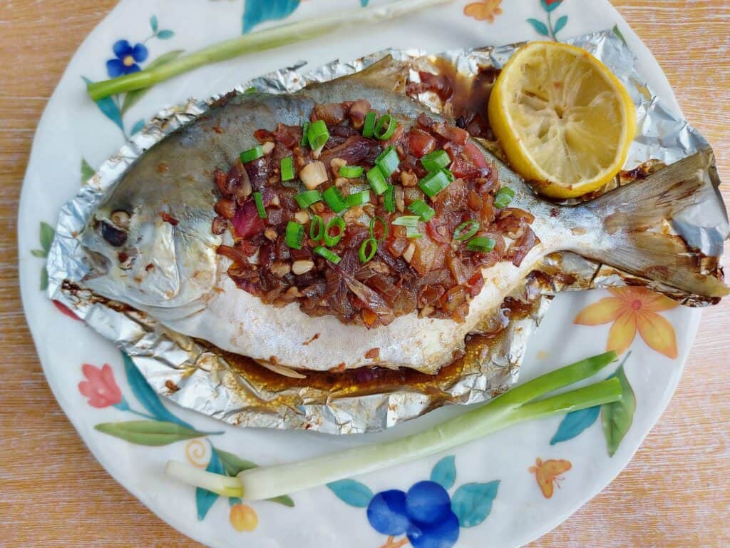 Whole baked pompano fish topped with onion-tomato soy sauce mixture with 2 onion leaves and 1 slice of lemon served on a plate