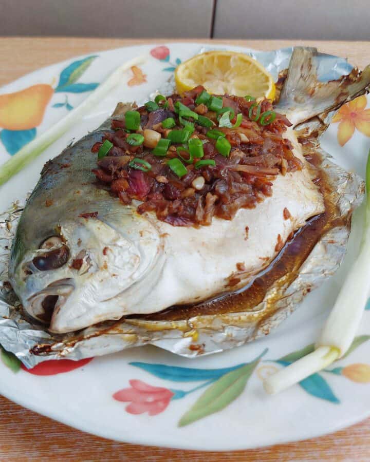 Whole baked pompano fish topped with onion-tomato soy sauce mixture with 2 onion leaves and 1 slice of lemon served on a plate