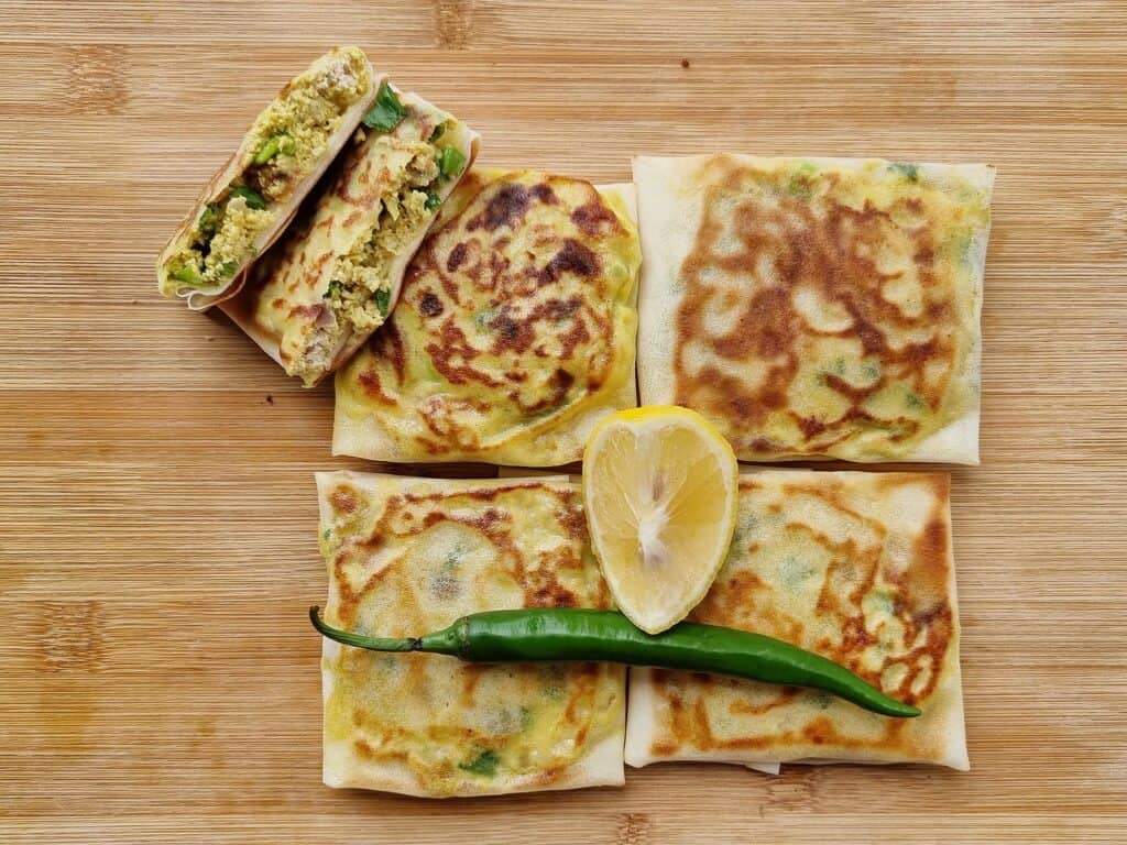 A golden Muttabaq pastry, layers of thin dough enveloping seasoned meat and greens, fried to crispy perfection in Saudi cuisine served with green chilies and lemon.