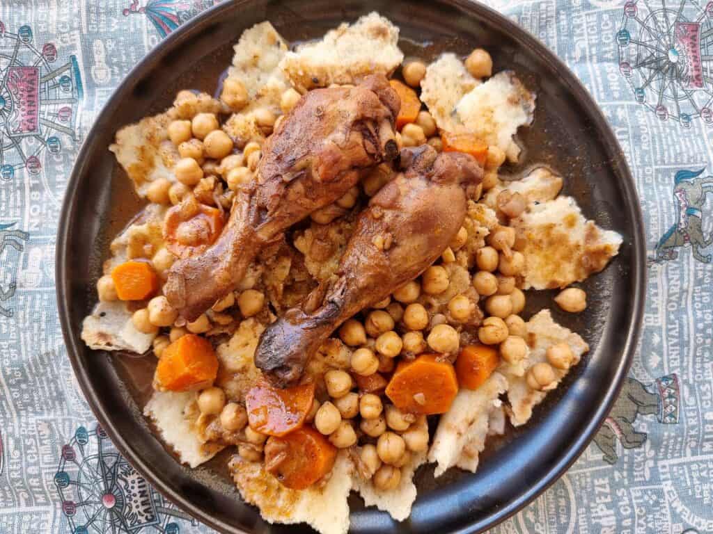 Chakhchoukha, a traditional Algerian dish, presents torn rougag flatbread soaked in a tomato-based broth with chicken, carrots, and chickpeas served on a plate